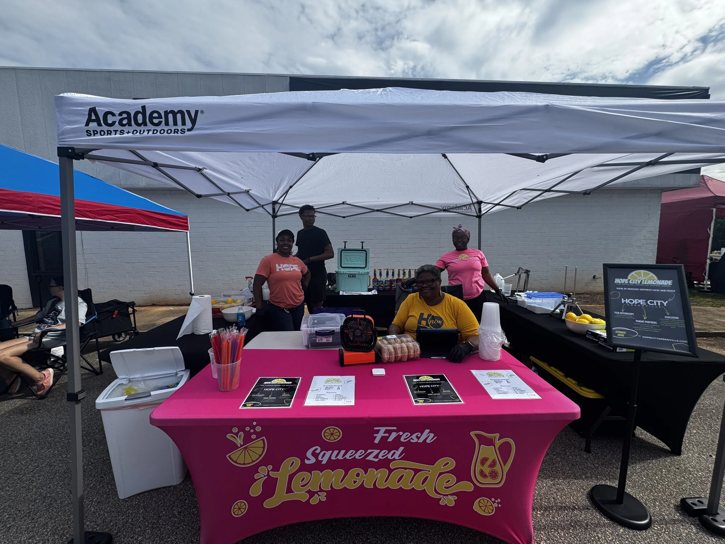 A group of outdoor charity event volunteers standing behind a pink lemonade stand with a black and white menu, under a white canopy with 'Academy Sports + Outdoors' branding, on a cloudy day.