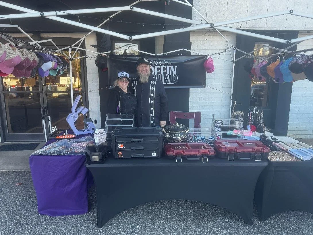 A man and woman standing behind a booth at an outdoor market with caps, jewelry, and various small items for sale.
