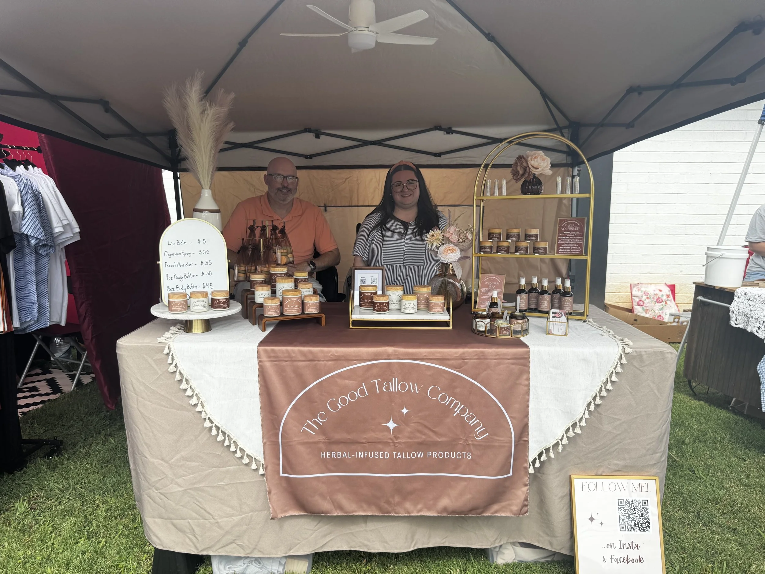 Display of herbal-infused all-natural skincare products at an outdoor market booth, including jars of balm, body butter, and sprays, with a man and woman behind the table, and a sign listing the products and prices. Tallow Butter company