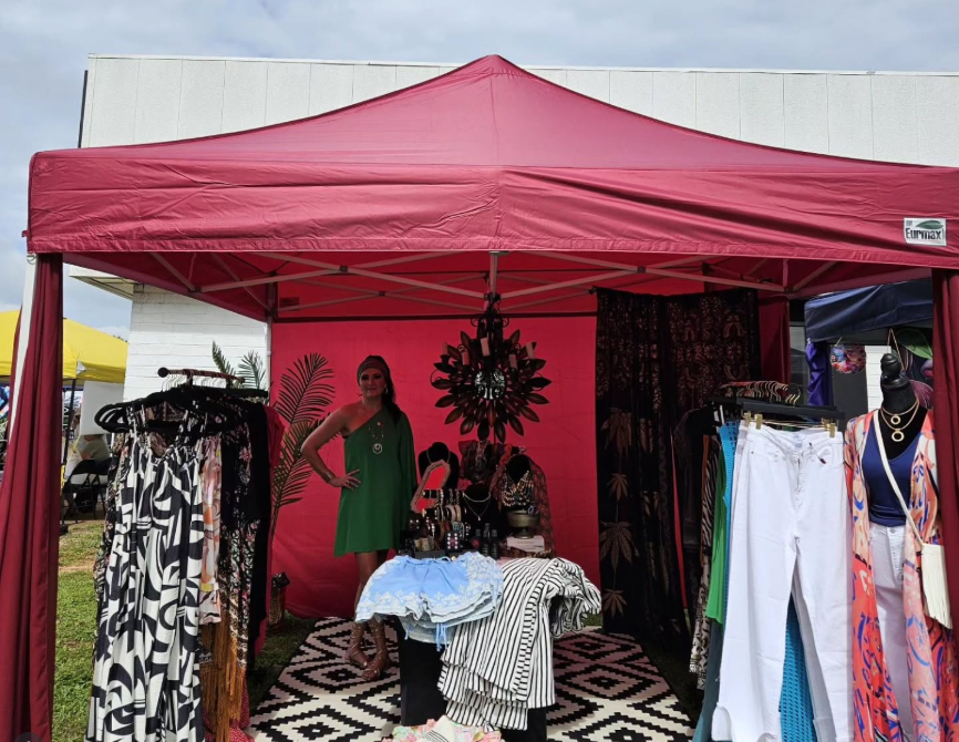 A woman in a green dress standing inside a vendor booth with clothing and accessories, under a red canopy at an outdoor market.