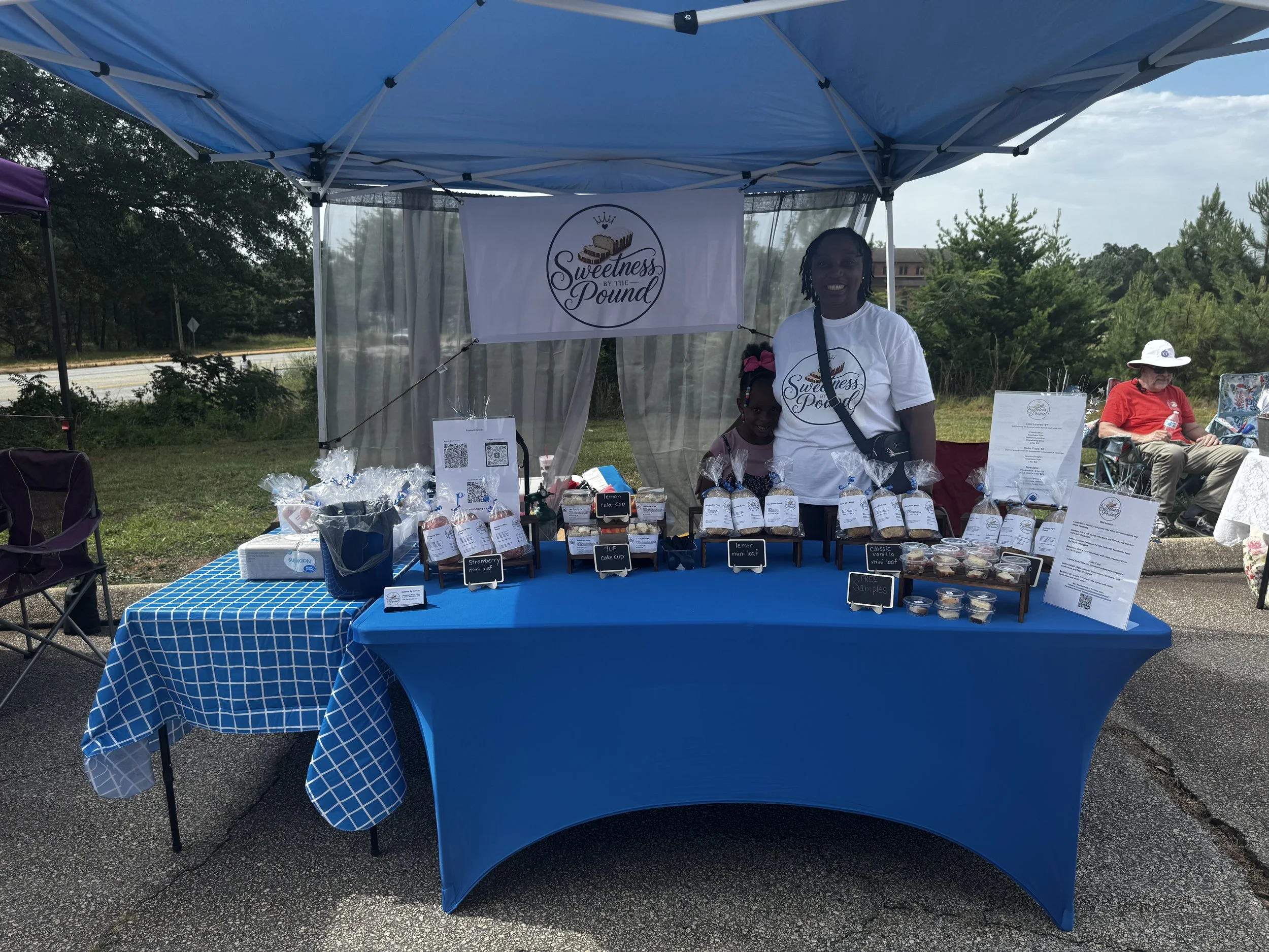 A woman and a young girl standing inside a blue canopy tent at an outdoor event, with a table displaying baked goods like lemon cake cups, strawberry mini loaf, and classic mini loaf, and a banner reading 'Sweetness by the Pound' behind them.