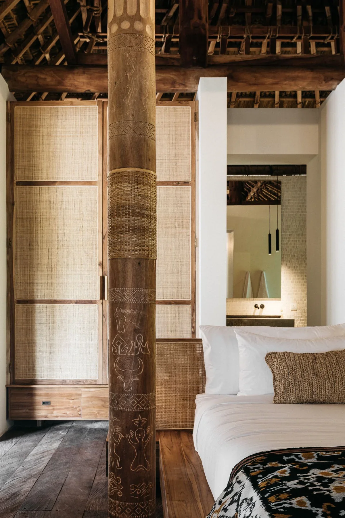 Interior of a bedroom with a white bed, beige pillows, wooden furniture, a carved wooden post with patterns, and a mirror in the background.
