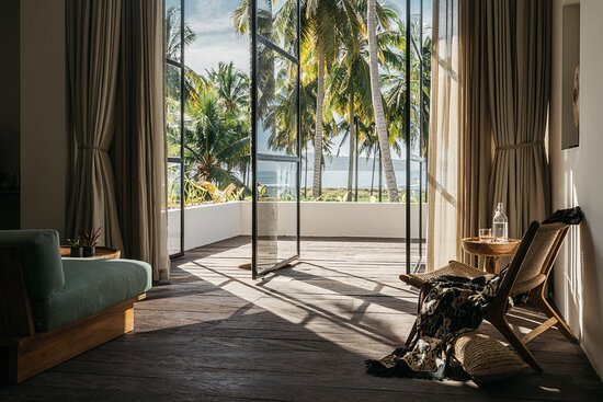 Room at Alamayah Sumba Yoga retreat with open glass doors leading to a balcony overlooking palm trees and the ocean, with curtains, a green sofa, and a chair with a water bottle on a small table.