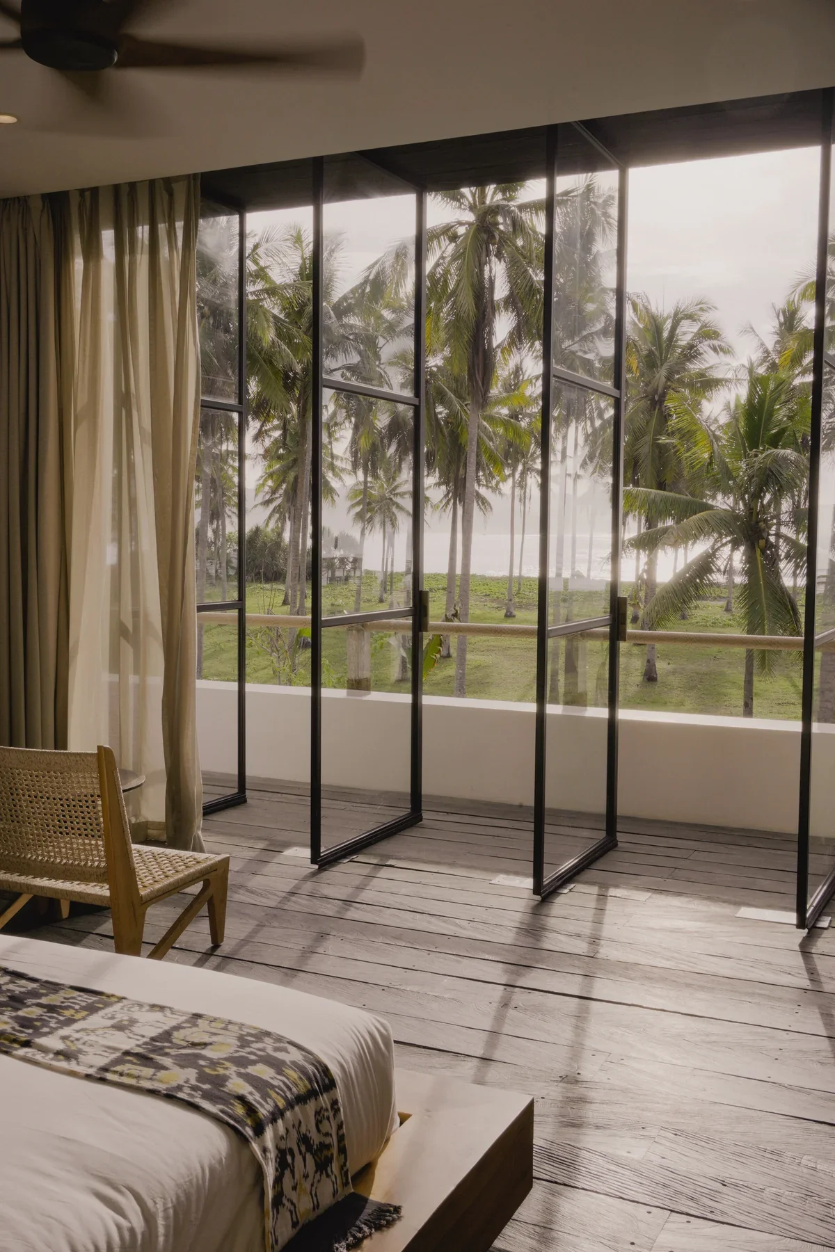 Beach view from a hotel room with large glass windows, white curtains, wooden floor, a chair, and a bed with a decorative blanket. Outside, palm trees are visible against a cloudy sky.