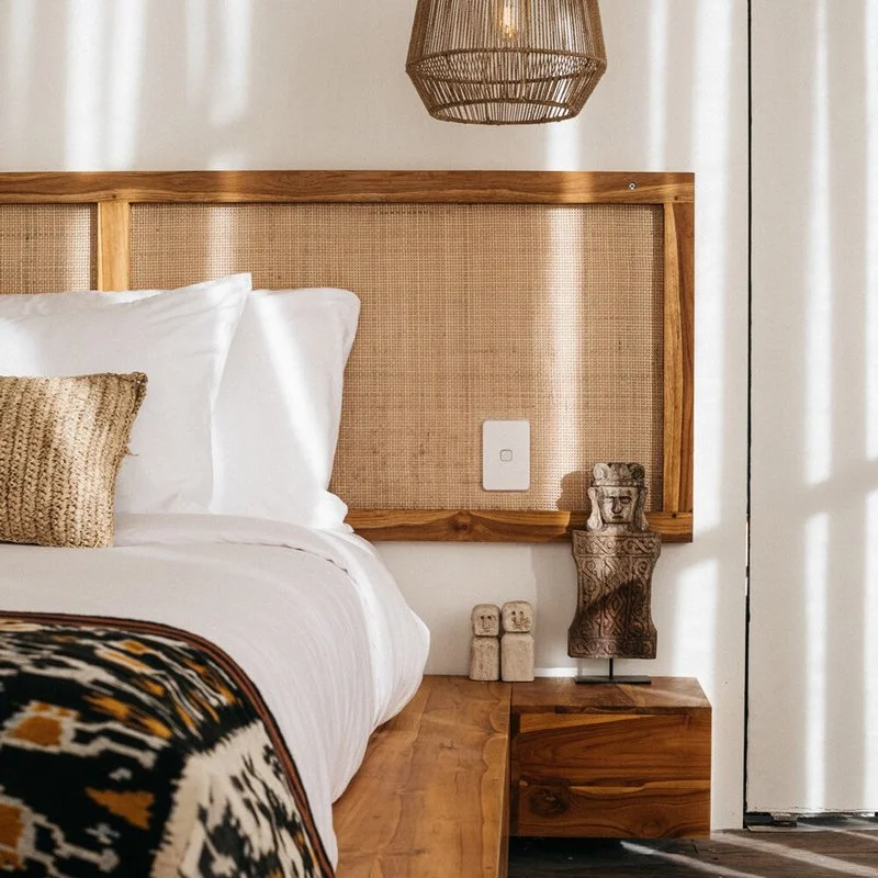 Bedroom with a bed,  a wooden headboard with woven panel, a wall lamp above, a small side table with sculptures, and natural light coming through a curtain at yoga and pilates retreat sumba indonesia
