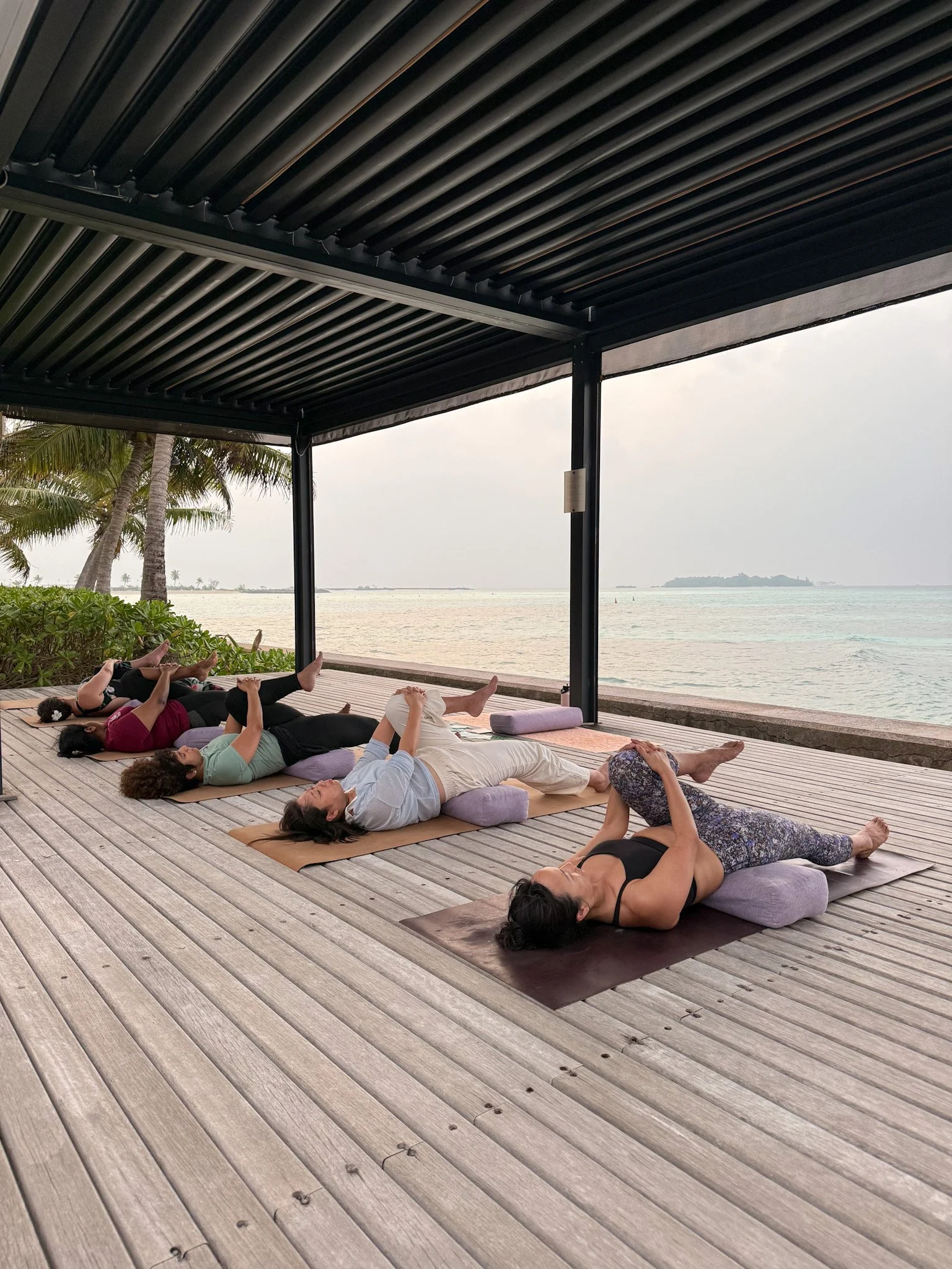 Yoga class on outdoor mat platform overlooking ocean at Bali pilates and yoga retreat with palm tree shaded open air studio
