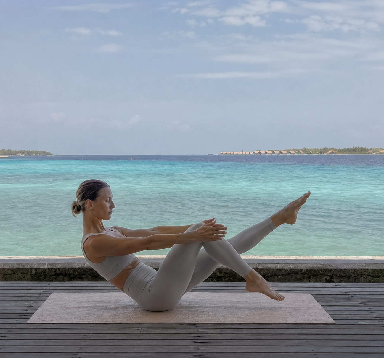 A woman practicing pilates in the Maldives at the yoga retreat on a mat outdoors by the water, with a view of a blue ocean and a partly cloudy sky.