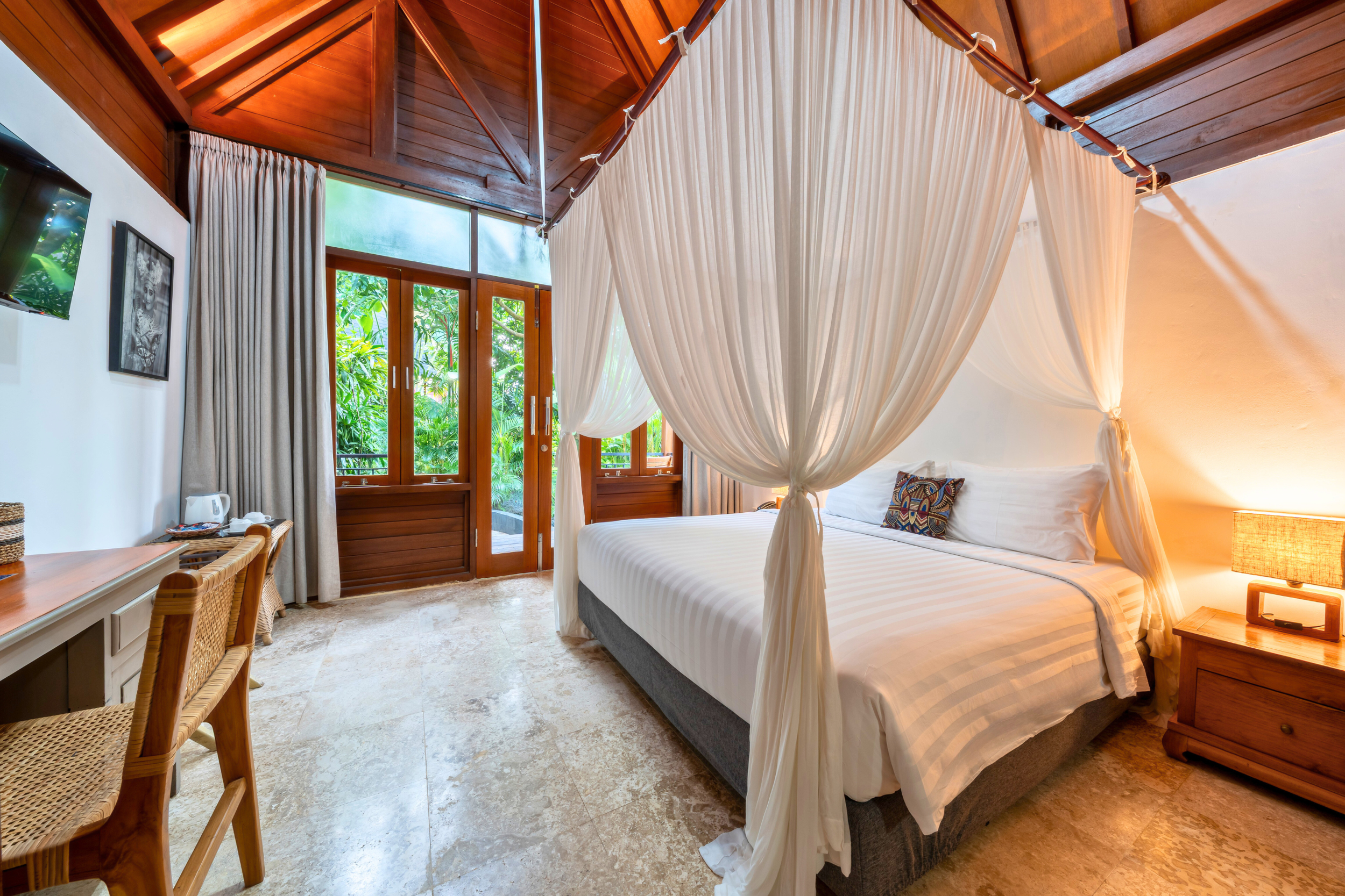Interior of a tropical hotel room with a four-poster bed and white curtains, wooden ceiling, large glass windows, and tropical greenery outside.