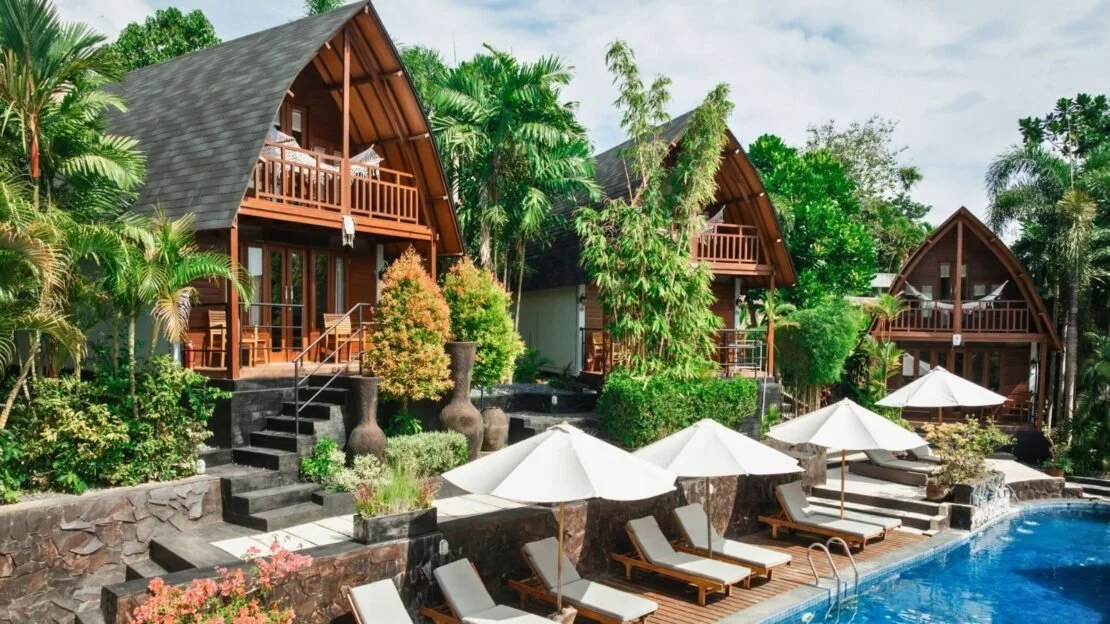 Resort with bungalows, swimming pool, lounge chairs, umbrellas, and lush greenery.
