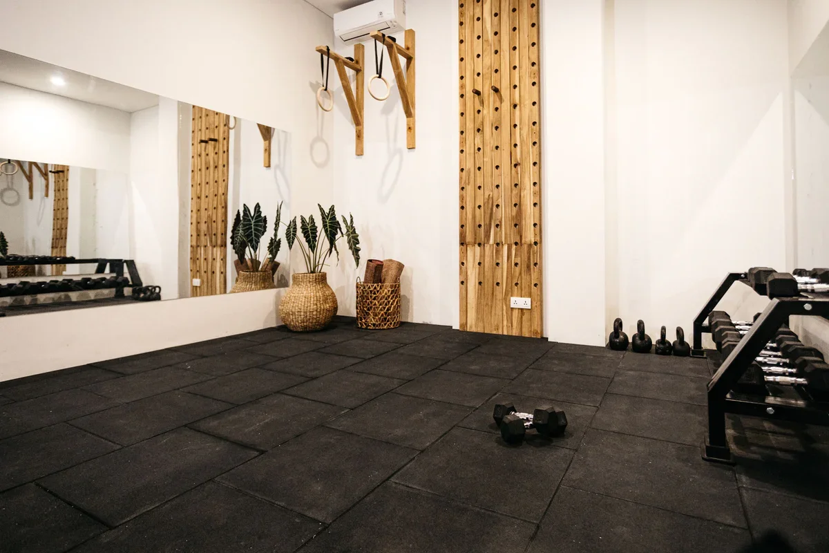 Home gym at Alamayah Sumba with black rubber flooring, dumbbells, kettlebells, a weight rack, gymnastic rings, a large mirror, decorative potted plants, and a wooden climbing wall panel.