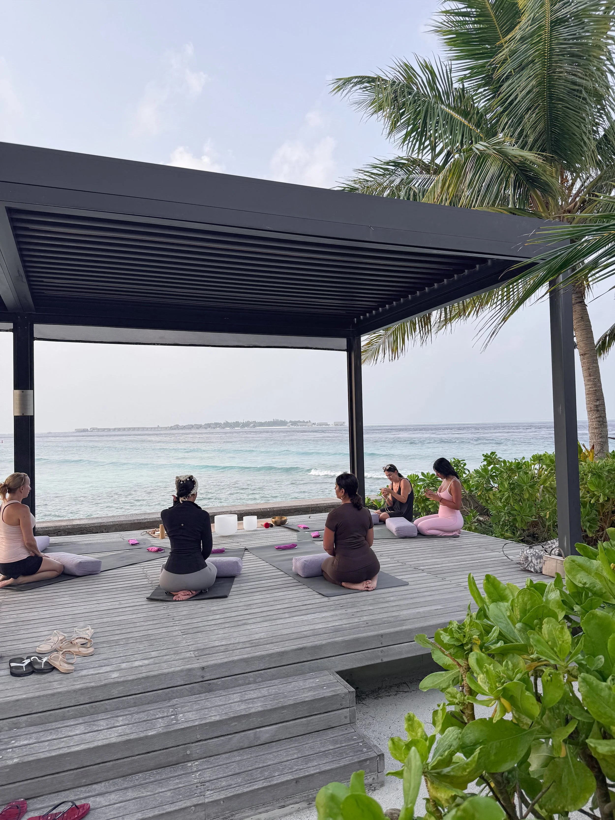People practicing yoga at a Maldives retreat on a wooden deck overlooking the beach with ocean waves, palm trees, and a partly cloudy sky.