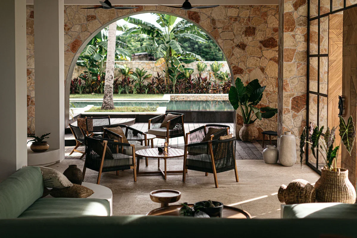 Indoor seating area with a view of a tropical garden through an arched opening, featuring wooden chairs, tables, large potted plants, and a stone wall.