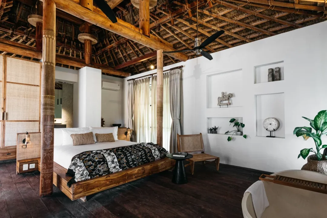 A cozy bedroom with a rustic wooden four-poster bed, white bedding, and decorative pillows. The room features dark hardwood floors, built-in wall shelves with decorative items, a large window with curtains, and a ceiling fan. There is also a small wo