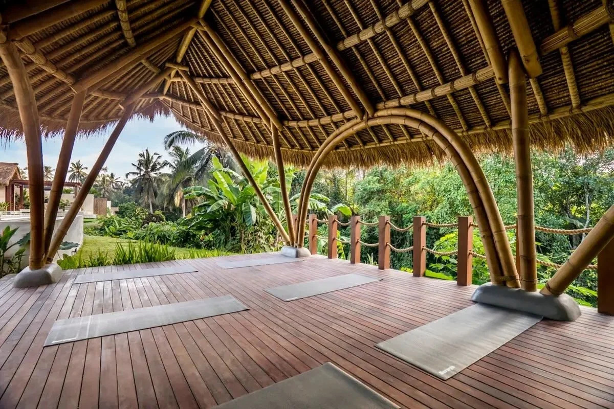 Open-air bamboo yoga studio with yoga mats on wooden floor, lush green tropical plants and trees outside, and a thatched roof overhead.