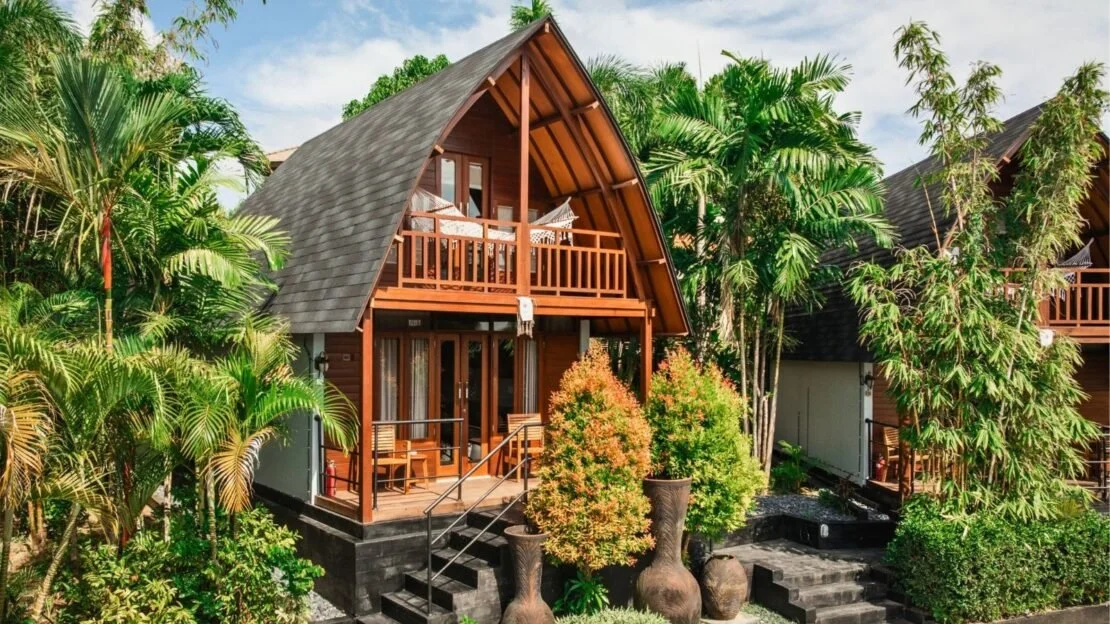 A wooden house with an A-frame roof, surrounded by tropical trees and plants, with a balcony and front porch.