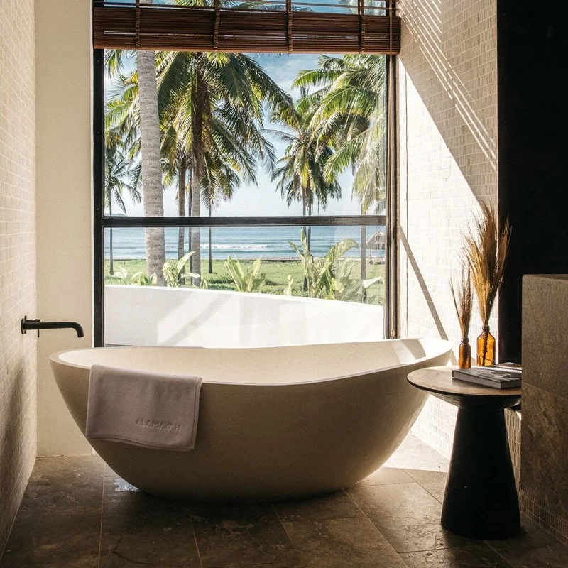 A modern bathroom with a freestanding bathtub facing a large window overlooking a tropical beach with palm trees and ocean view.