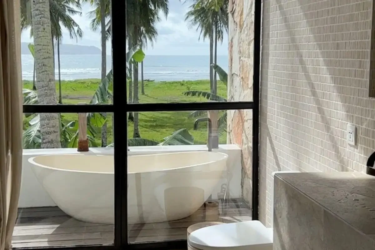 A freestanding bathtub on a balcony  at Alamayah Sumba Yoga & Pilates retreat overlooking a tropical beach with palm trees and ocean in the background, seen through a glass window.