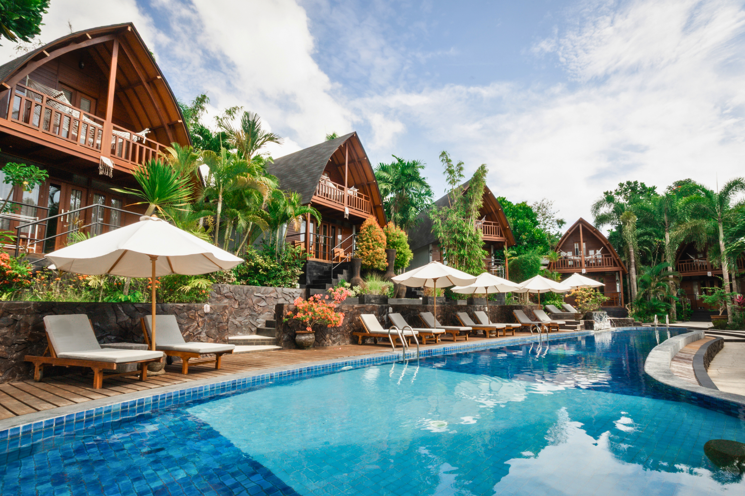 Resort with wooden A-frame cabins, swimming pool with lounge chairs and umbrellas, lush green trees, and a partly cloudy sky.