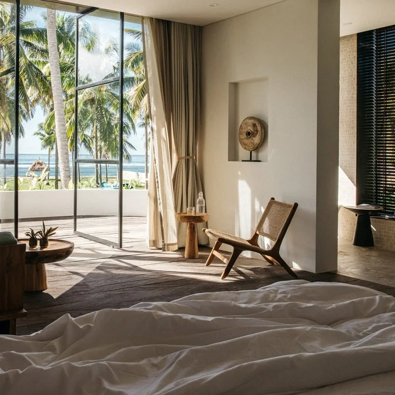 Beachfront hotel room with large glass doors opening to a patio, with palm trees and ocean view outside, beige curtains, a wicker chair, and a bed with white linens.