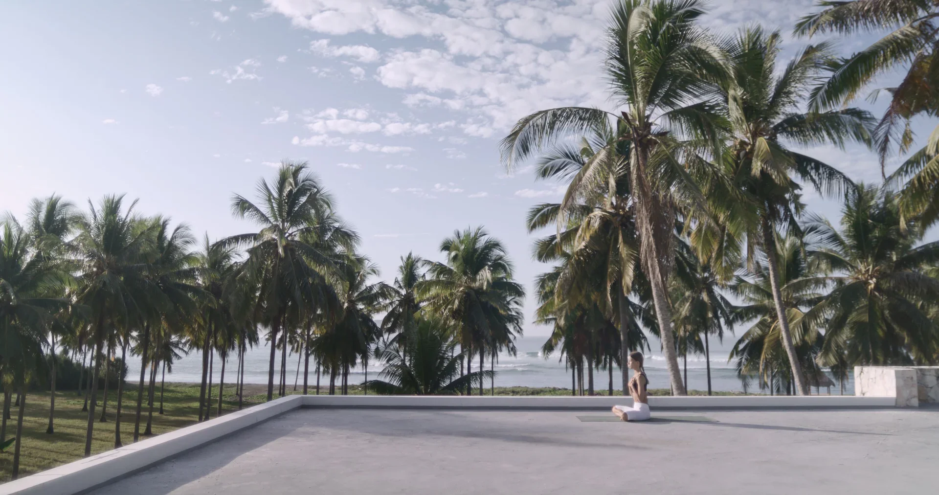 A woman practicing yoga in a seated meditation pose outdoors on a concrete surface, surrounded by a tropical landscape with tall palm trees and the ocean in the background on a sunny day.