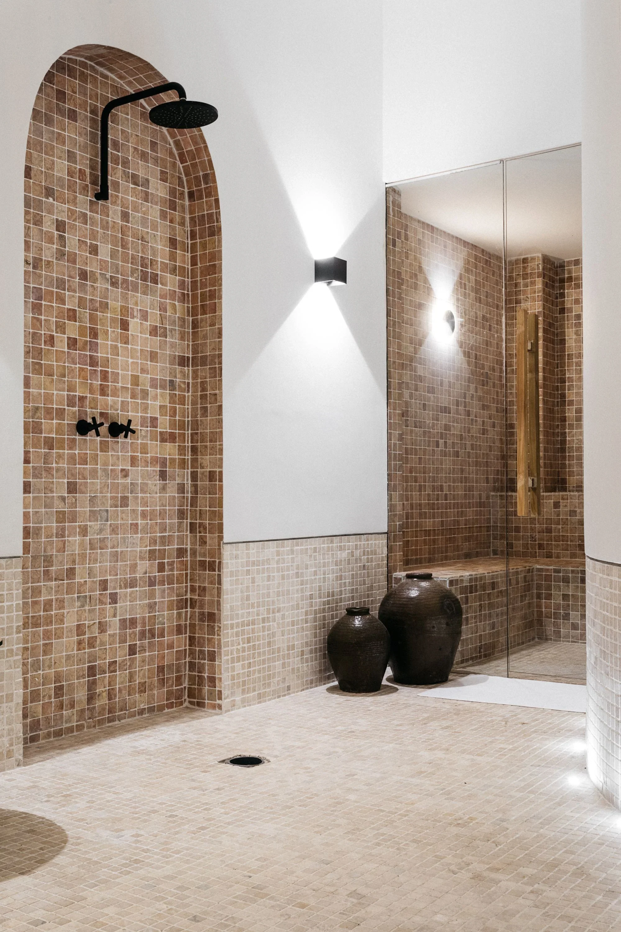 Modern bathroom with beige mosaic tiles, black showerhead, matching faucets, decorative black vases, and a large mirror.