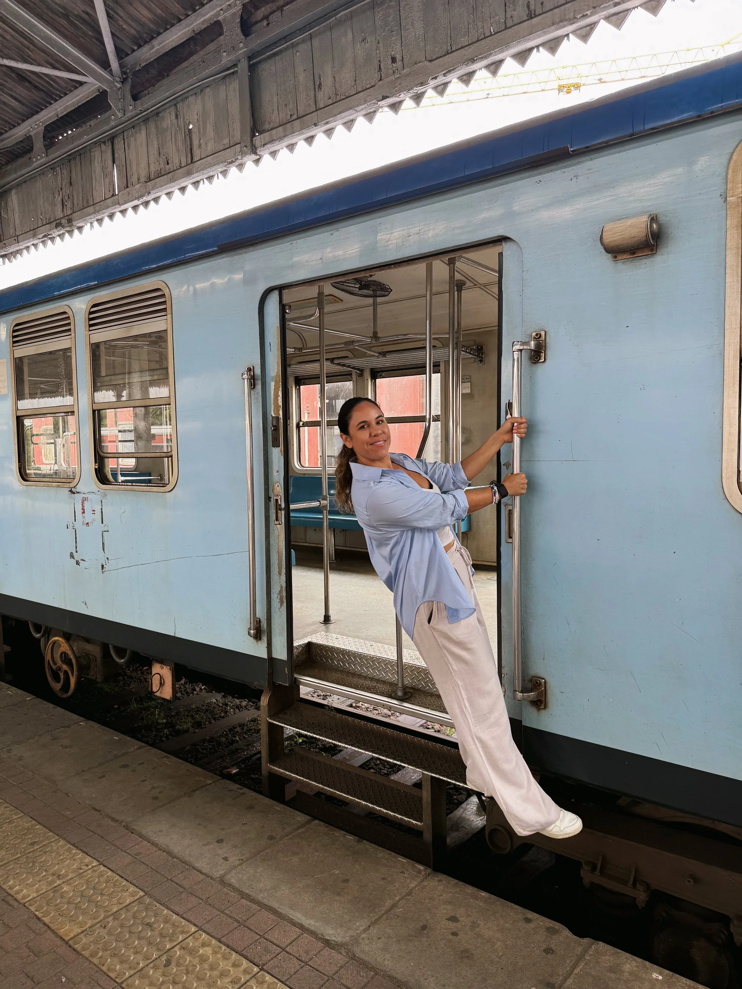 A woman smiling and holding onto a train door handle while standing on the train's exterior platform, next to a vintage blue train car.