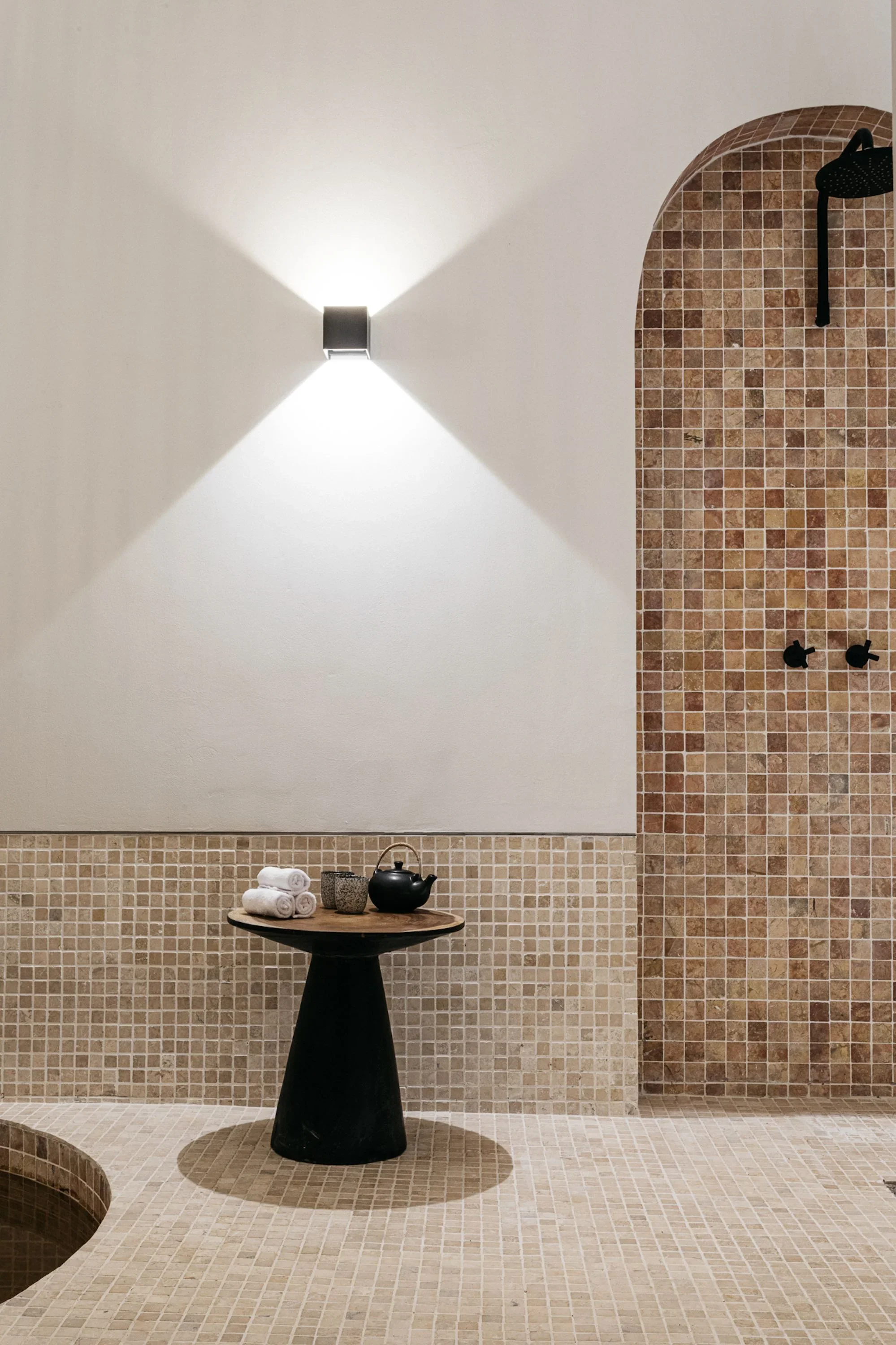 A spa or steam room with a beige tiled floor and wall, a black pedestal table with rolled white towels and a tea set, and a wall-mounted black showerhead. The wall features a white section with a modern wall light casting a triangular light pattern, 