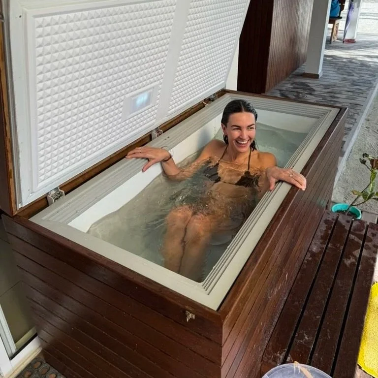 A woman relaxing in a hot tub with a lid open, sitting on the edge and smiling.
