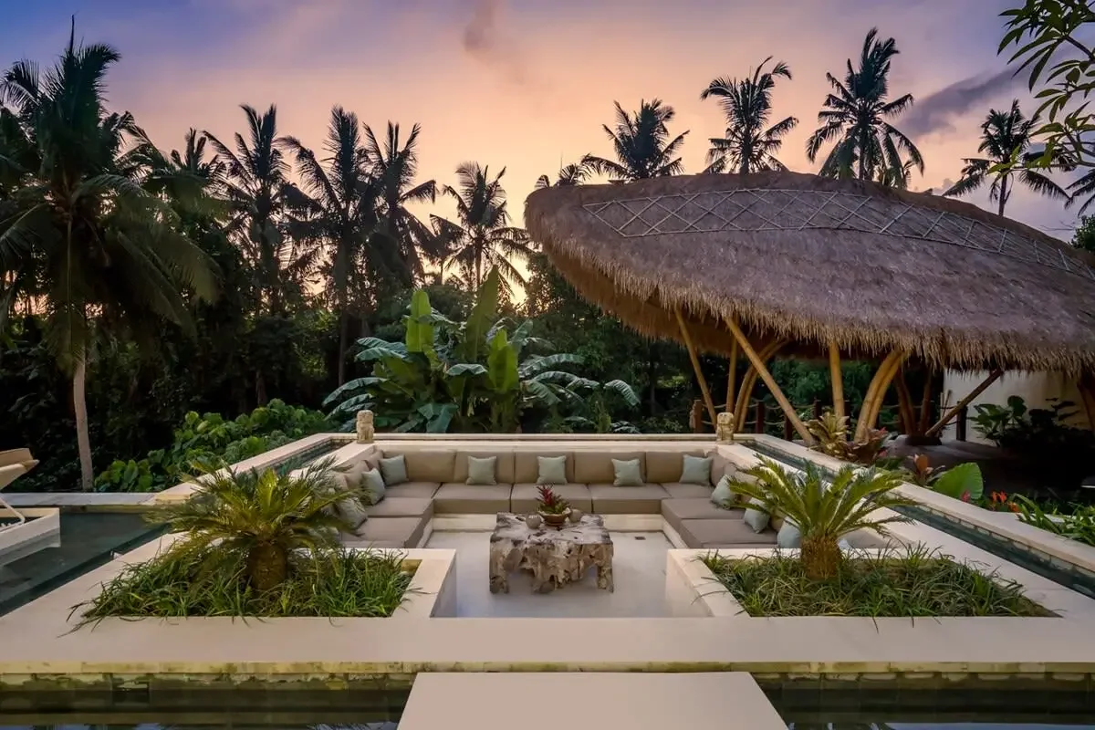 A luxurious outdoor patio with a large L-shaped sectional sofa, small palm trees in planters, and a thatched roof tiki hut, set against a lush tropical backdrop of palm trees and sunset sky.