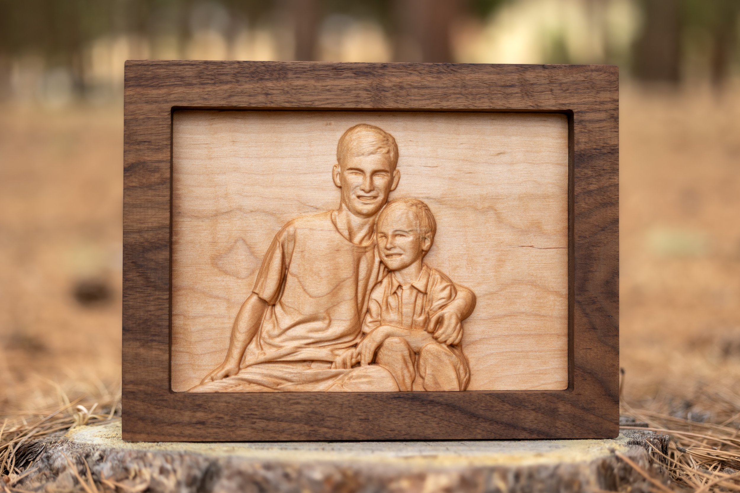 A wooden carved picture of two young boys, one older with short hair and the other younger with longer hair, both smiling, framed in a wooden border, set outdoors among pine needles and trees.