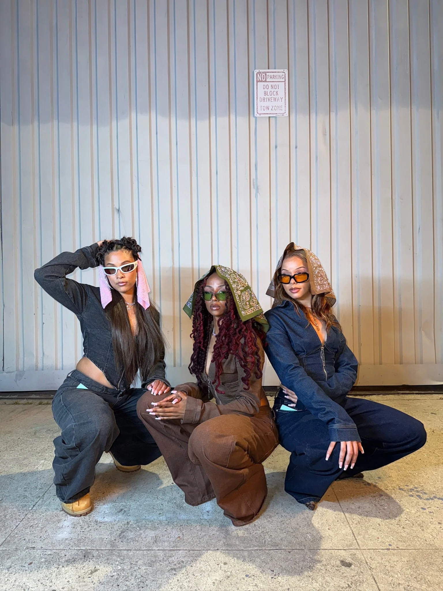 Four women posing in front of a metal wall, wearing sunglasses and hooded jackets, with one woman sitting on a cardboard box.