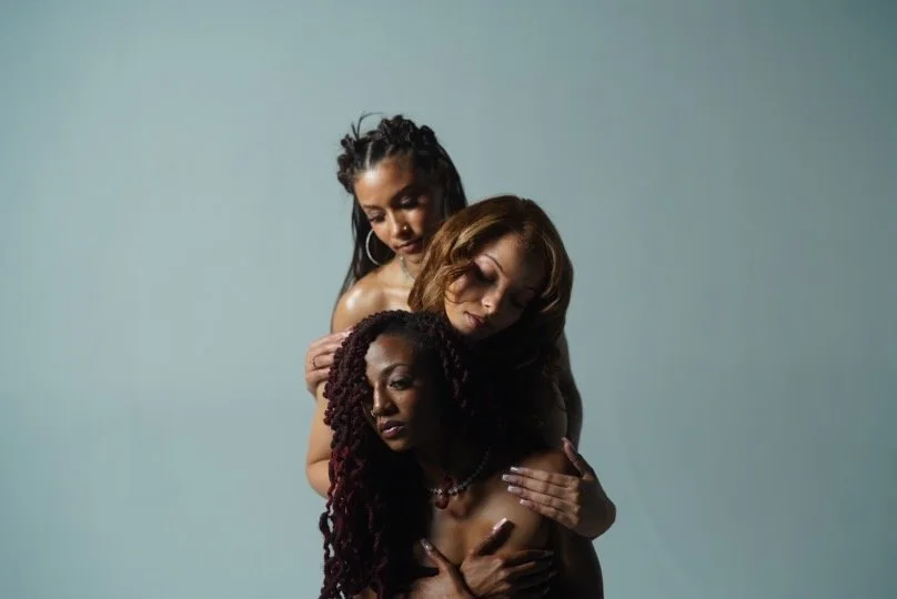 Three women posing in an artistic portrait with a plain background, one with braids, one with curly hair, and one with straight hair.