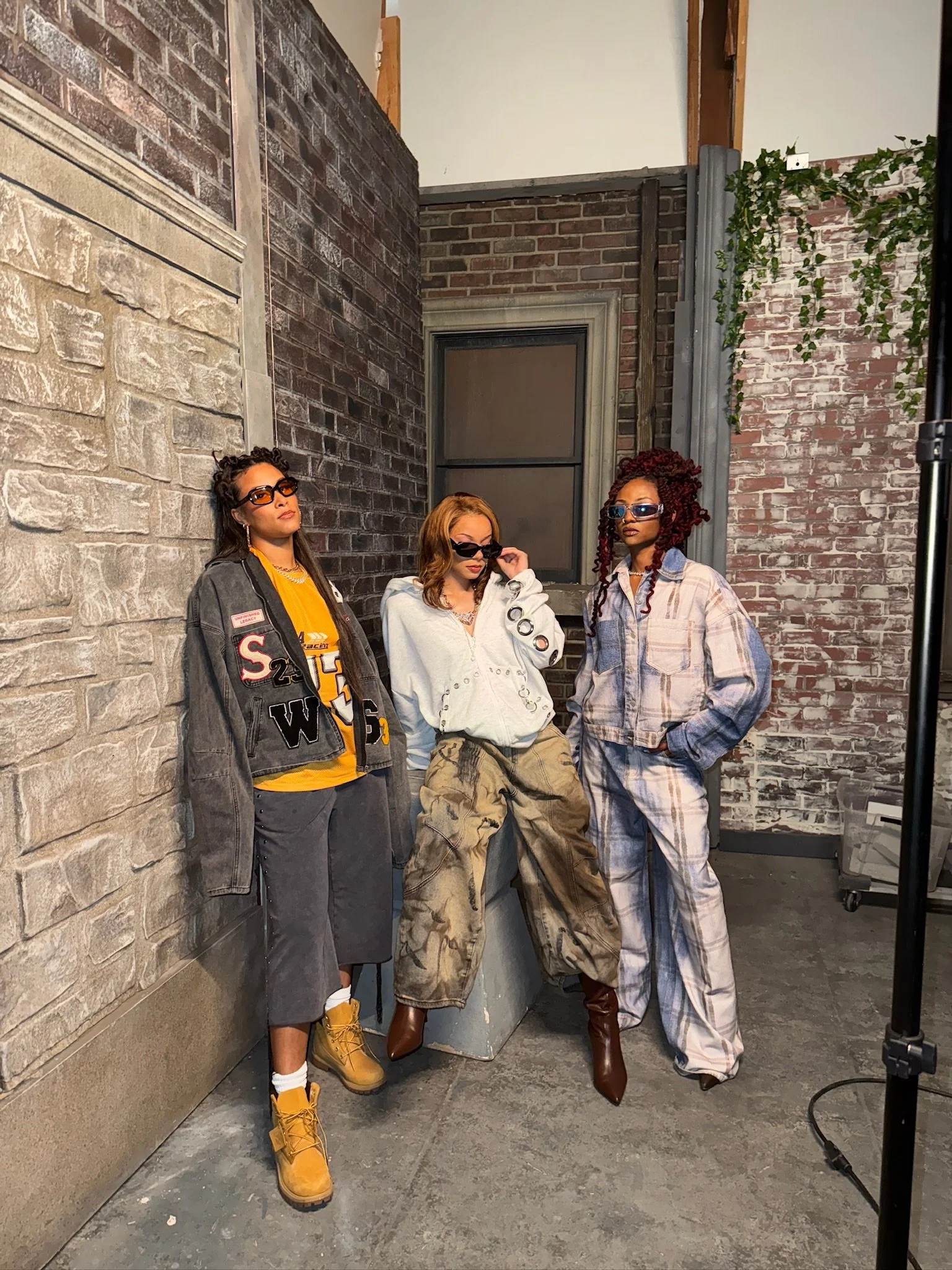 Four women standing in an industrial-style room with brick walls and concrete floor, wearing casual and streetwear outfits, sunglasses, and boots.
