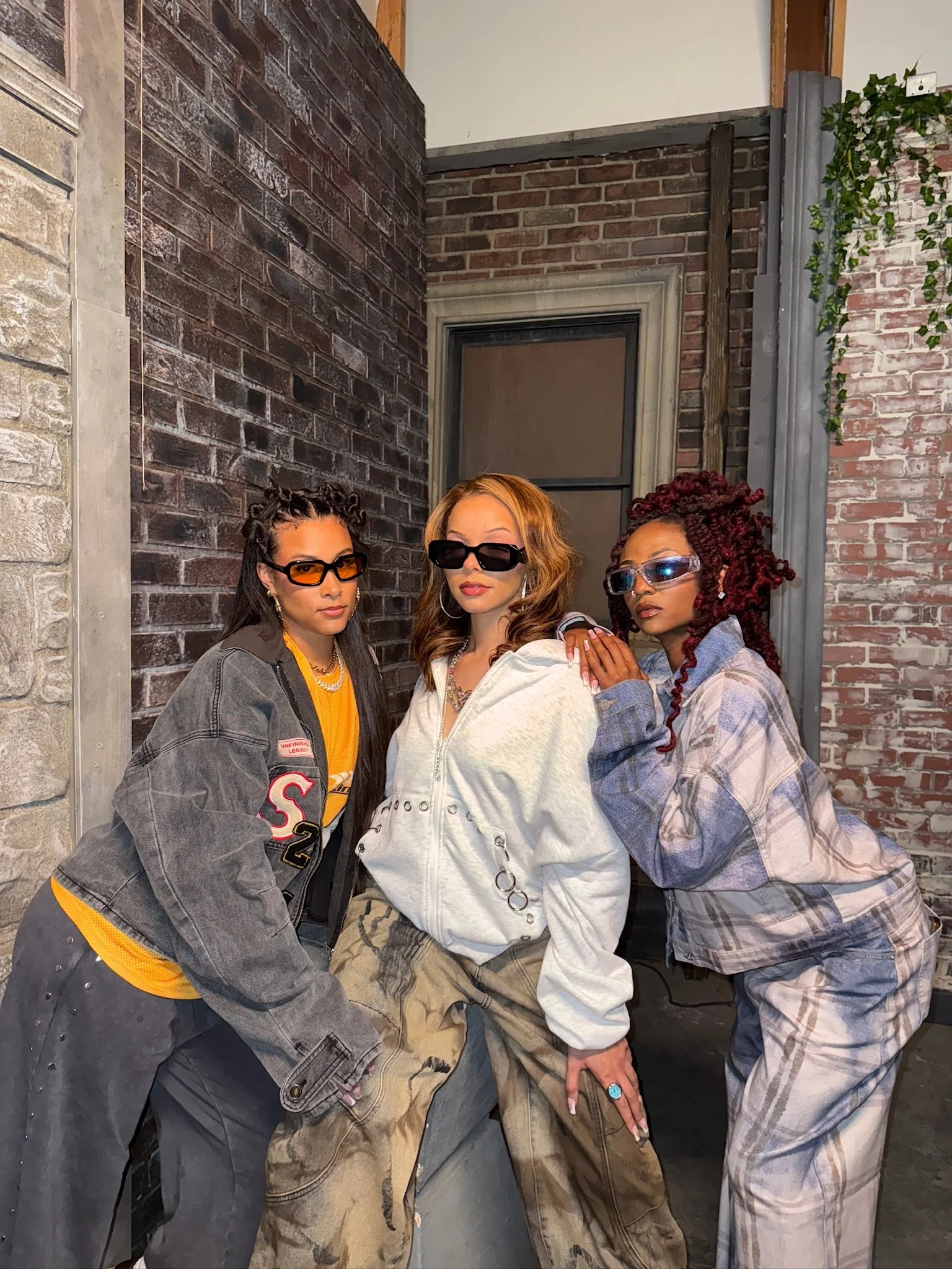 Three women with distinct hairstyles and sunglasses posing together outdoors against a brick wall.