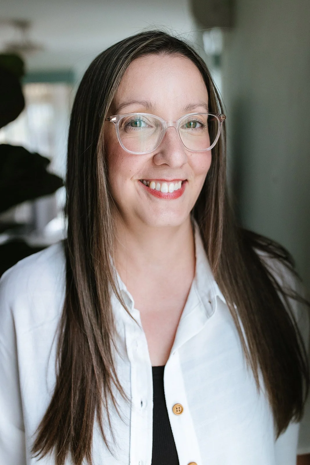A woman with long brown hair, wearing clear glasses, a white shirt, and a black top, smiling indoors.