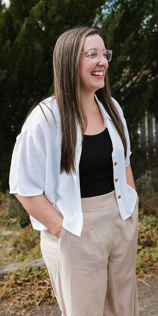 Elise Heerde - A woman with long brown hair and glasses laughing outdoors, wearing a white short-sleeved shirt over a black tank top and beige pants.