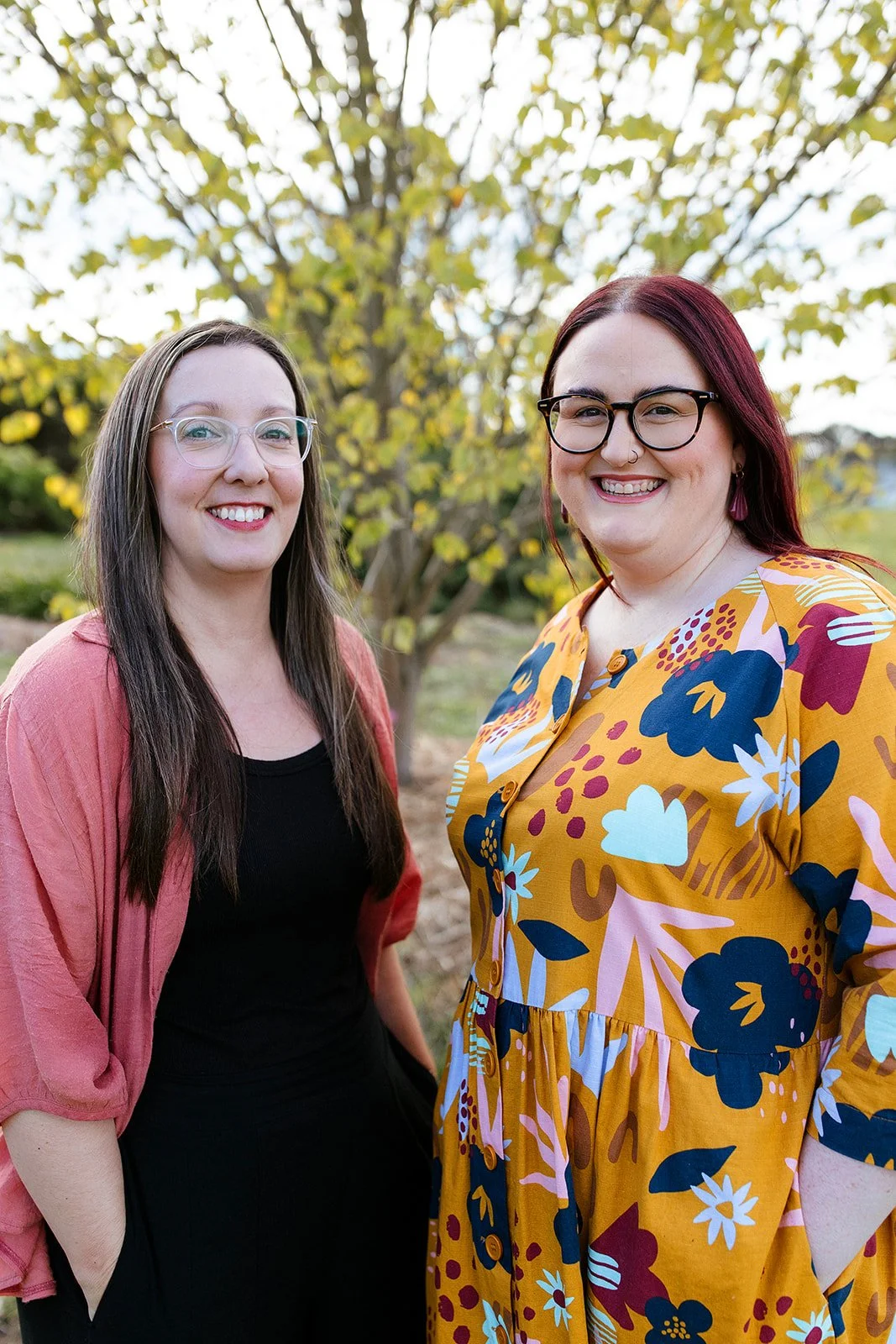 Elise Heerde and Sam Sellers - Two women smiling outdoors near a tree with yellow and green leaves, one wearing a black top and pink cardigan, the other wearing a colorful, floral-patterned dress and glasses.