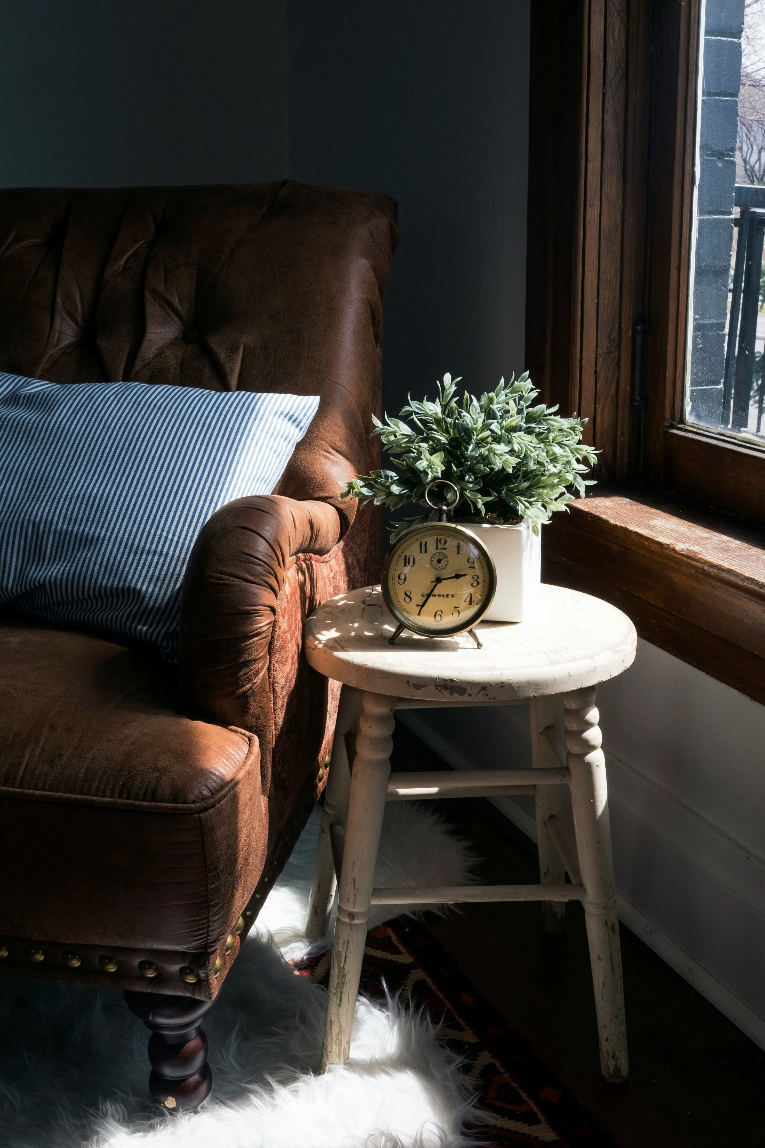 A cozy corner with a brown leather armchair, a blue and white striped pillow, a white furry rug, a small white side table with a potted plant, an alarm clock, and a window showing a street view outside.