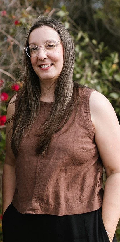 Elise Heerde - A woman with long brown hair, wearing glasses and a sleeveless brown top, smiling outdoors with greenery and pink flowers in the background.