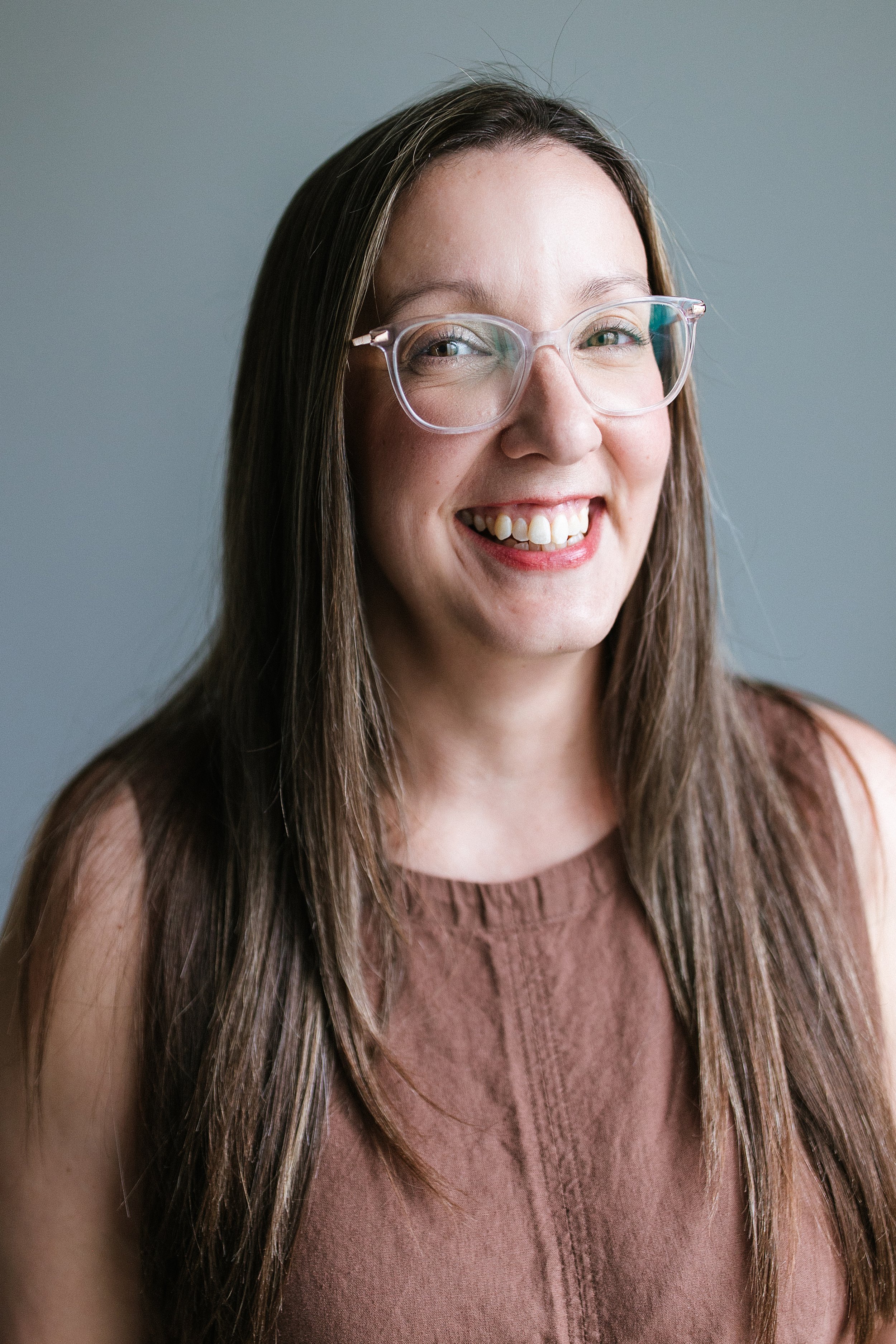 A woman with long brown hair and glasses smiling and looking slightly to the side, wearing a sleeveless brown top.