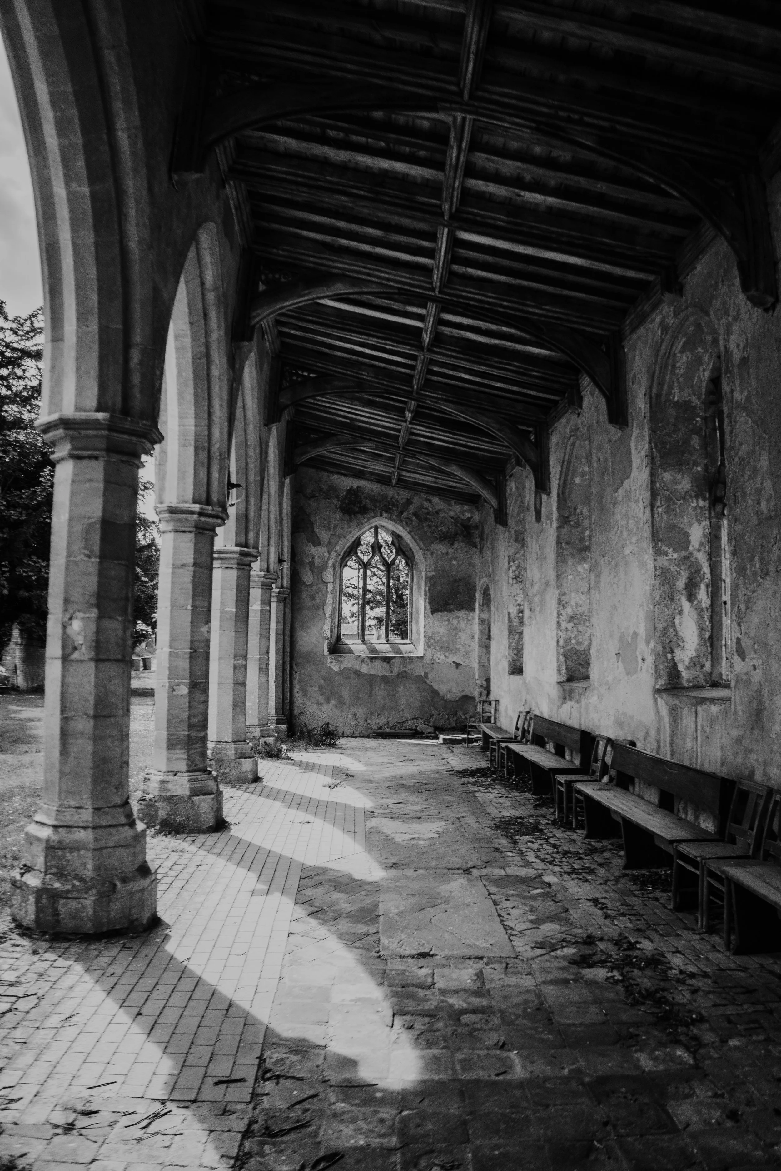 Black and white photo of an abandoned church or chapel with broken benches, peeling walls, and large arched windows. The wooden ceiling is supported by decorative stone columns.