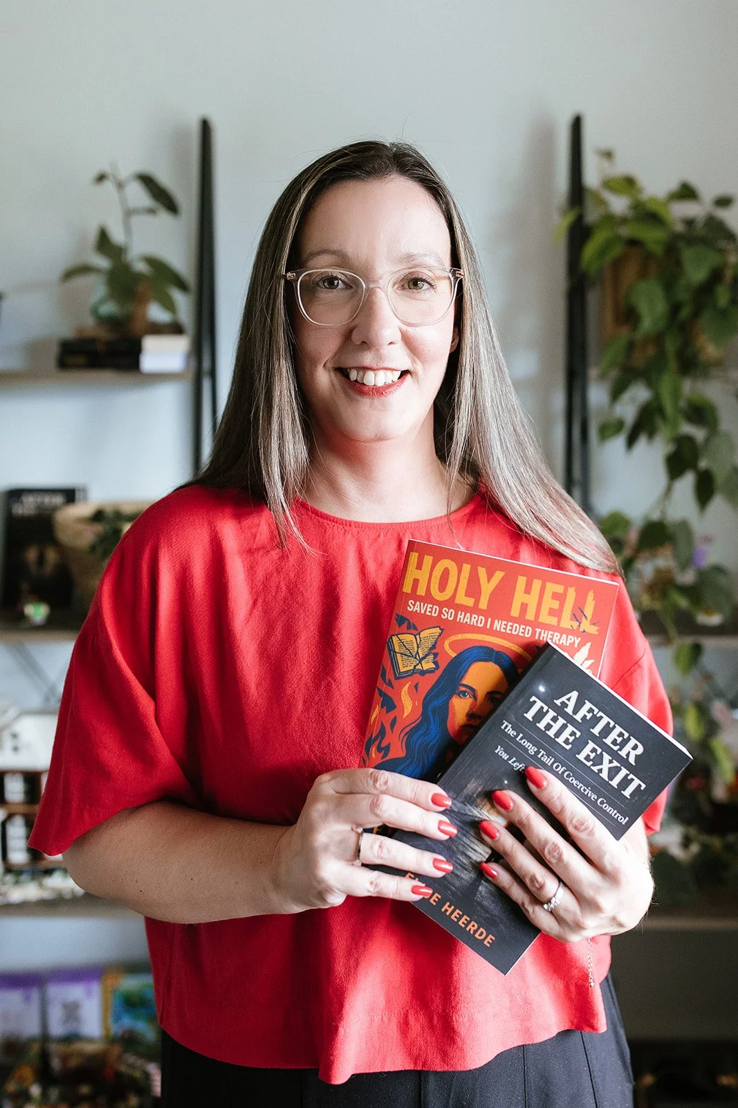 Elise Heerde - A smiling woman wearing glasses and a red shirt, holding two books titled "Holy Hell" and "After the Exit," standing in a room with shelves and plants in the background.