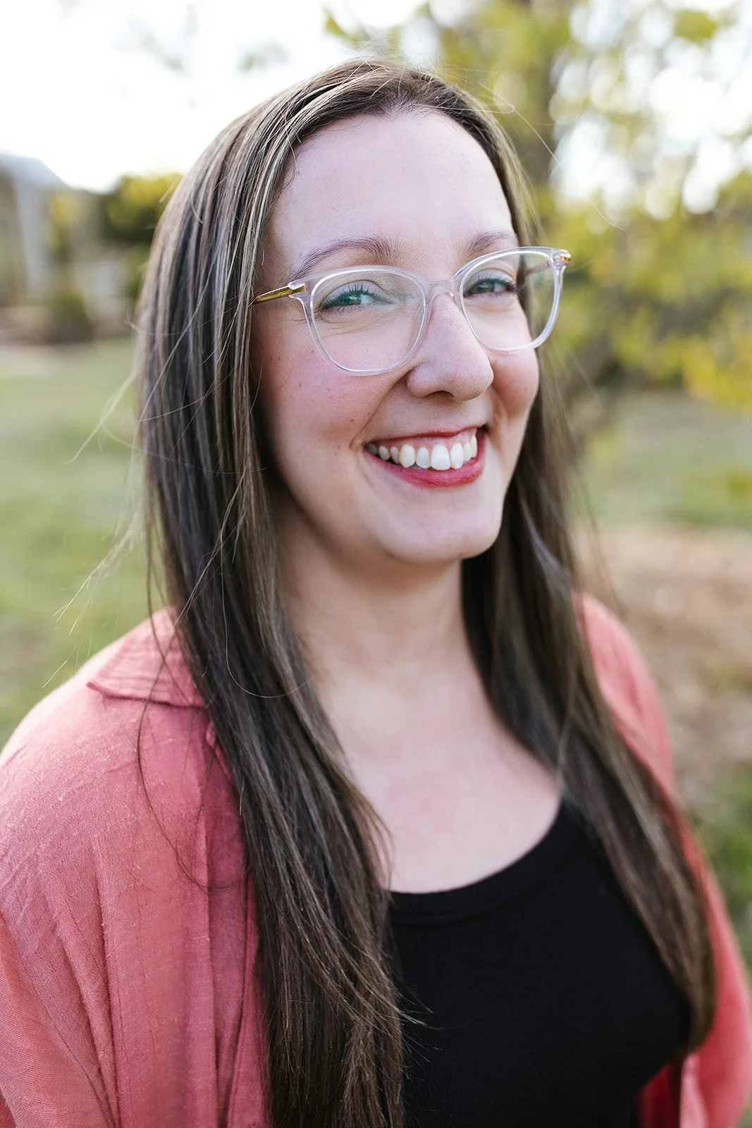 Elise Heerde - A young woman with long brown hair, wearing glasses, smiling outdoors with trees in the background.