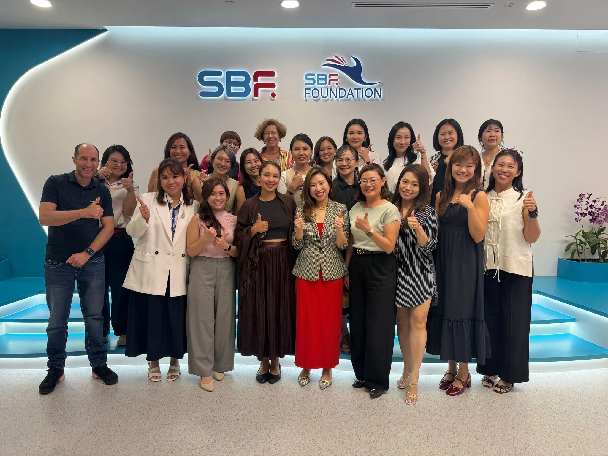 A group of around 20 diverse women and a man posing together in an indoor setting with a white wall in the background. The wall has logos that read 'SBF' and 'SBF Foundation' with a design of a hand symbol. The group is smiling, with some giving thum
