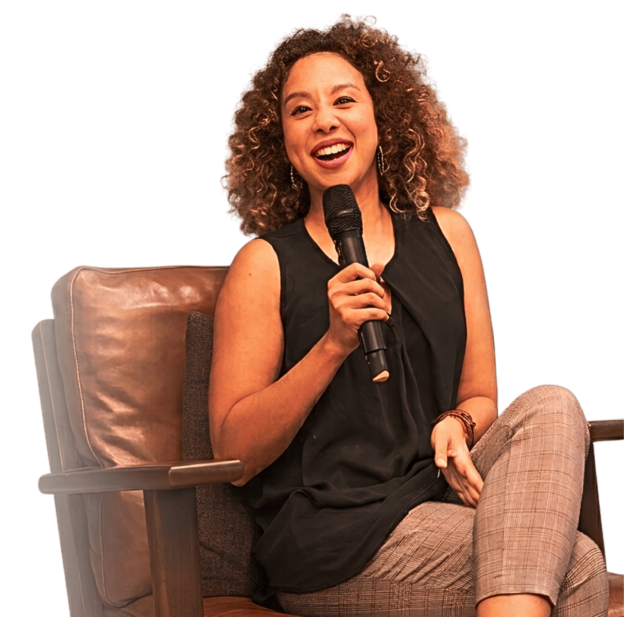 Woman with curly hair smiling and holding a microphone, sitting in a chair during a speaking event.
