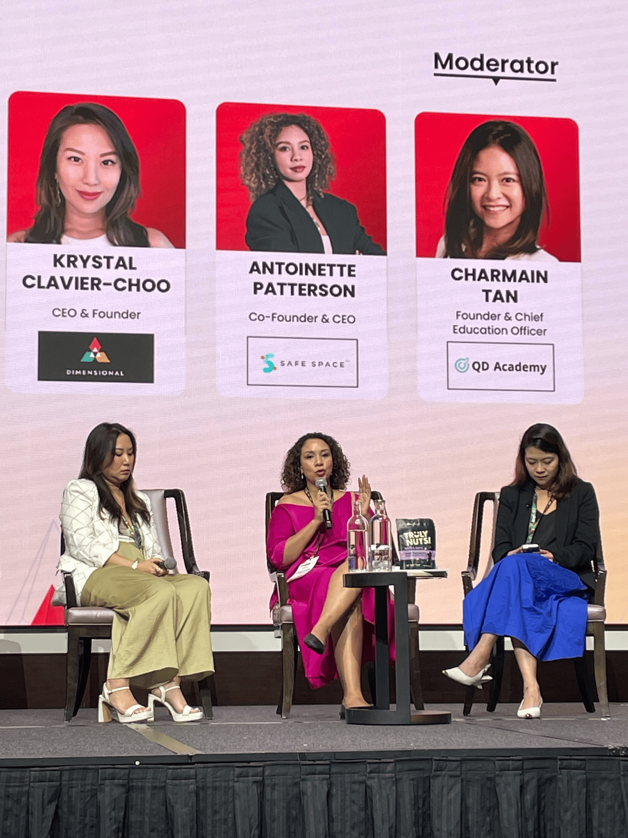 Three women sitting on stage at a panel discussion with a large screen behind them showing photos and titles. The women are dressed in formal or business attire, and the woman in the middle is speaking into a microphone.