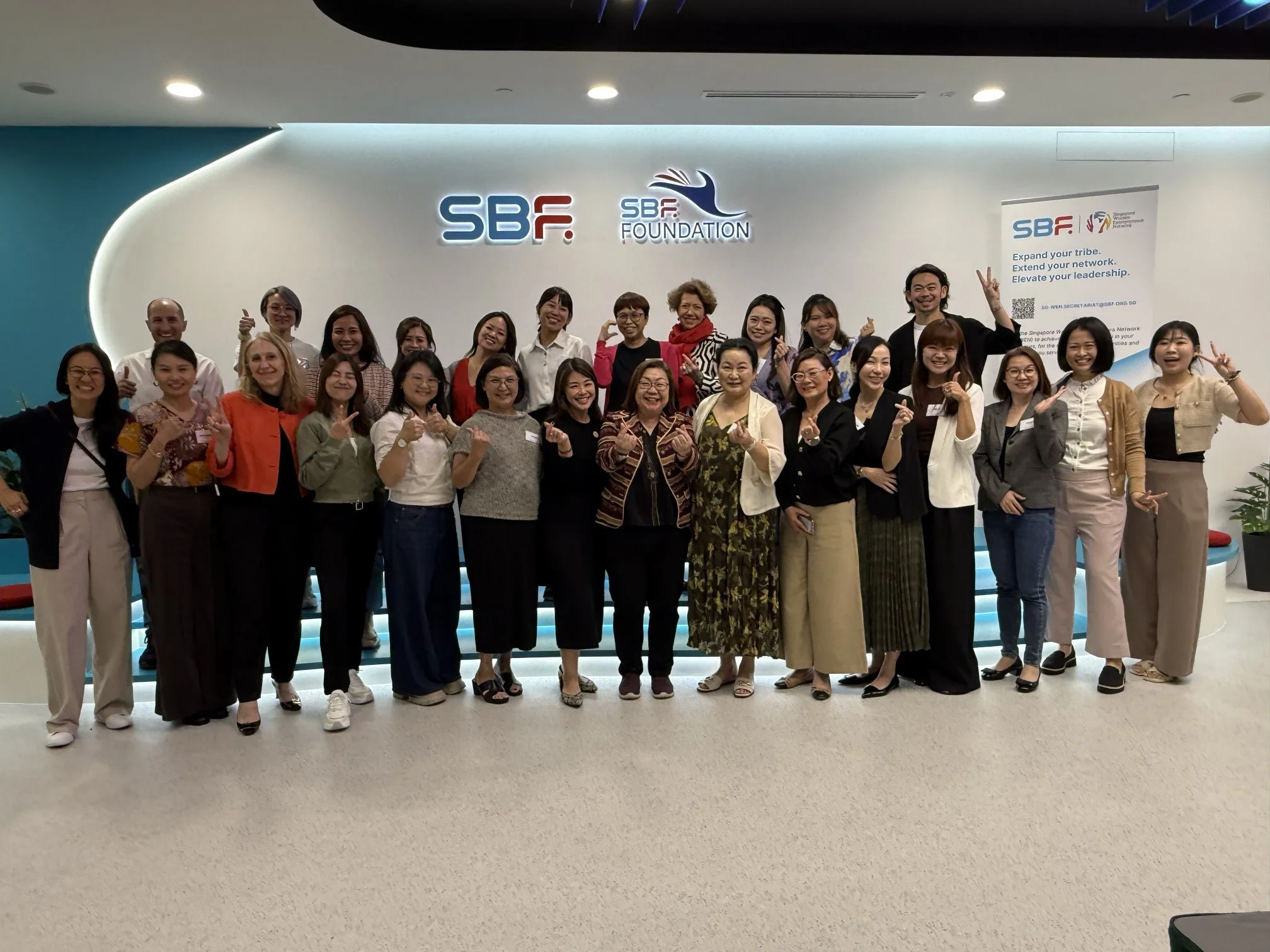 Group of diverse women and men in business casual attire standing on stairs against a wall with the logos of SBf and SBf Foundation, smiling and making hand gestures in a professional setting.