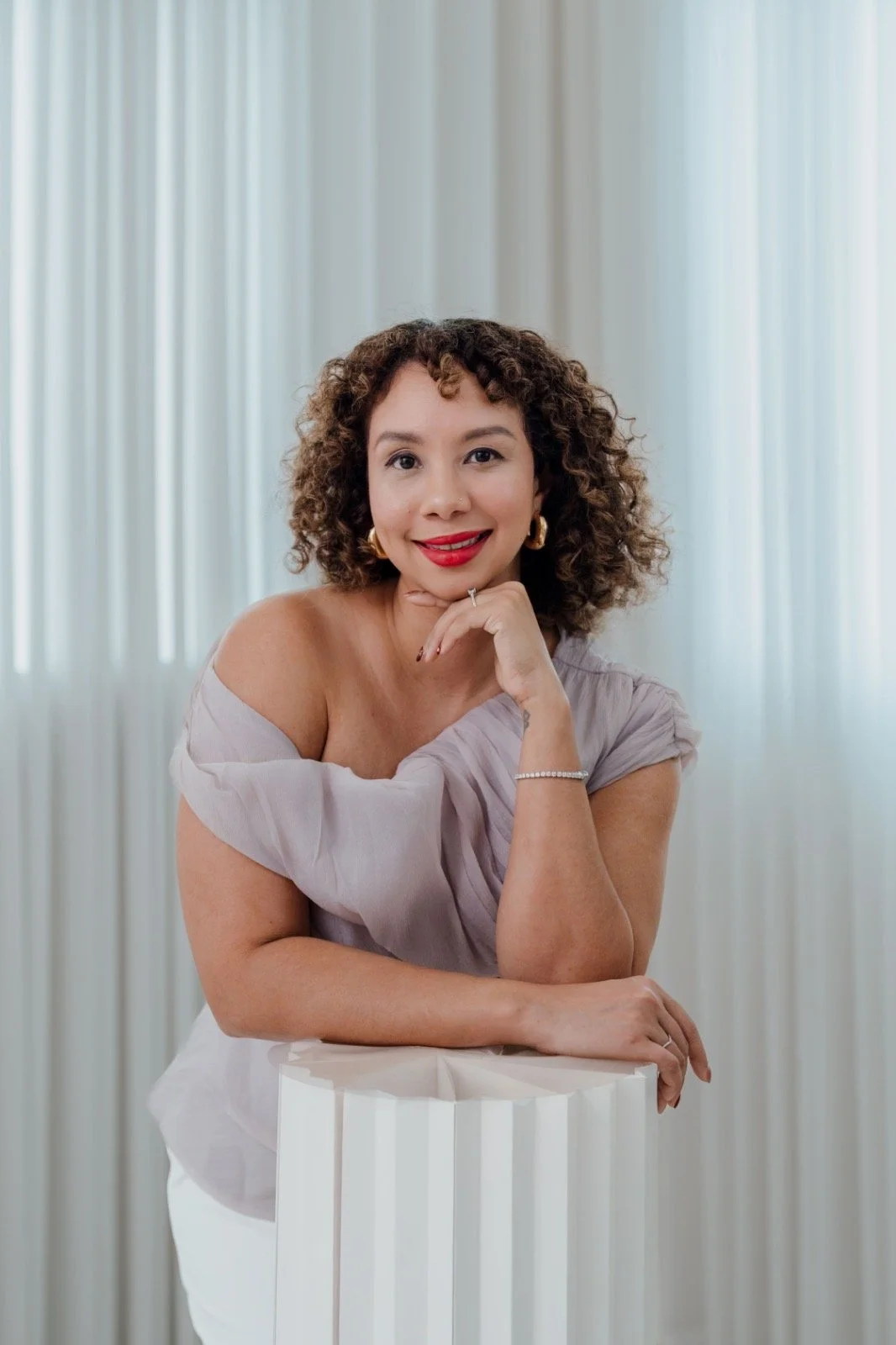Portrait of a woman with curly hair wearing a light-colored off-shoulder top, gold jewelry, and red lipstick, smiling and resting her chin on her hand, standing in front of a light-colored, vertical striped background.