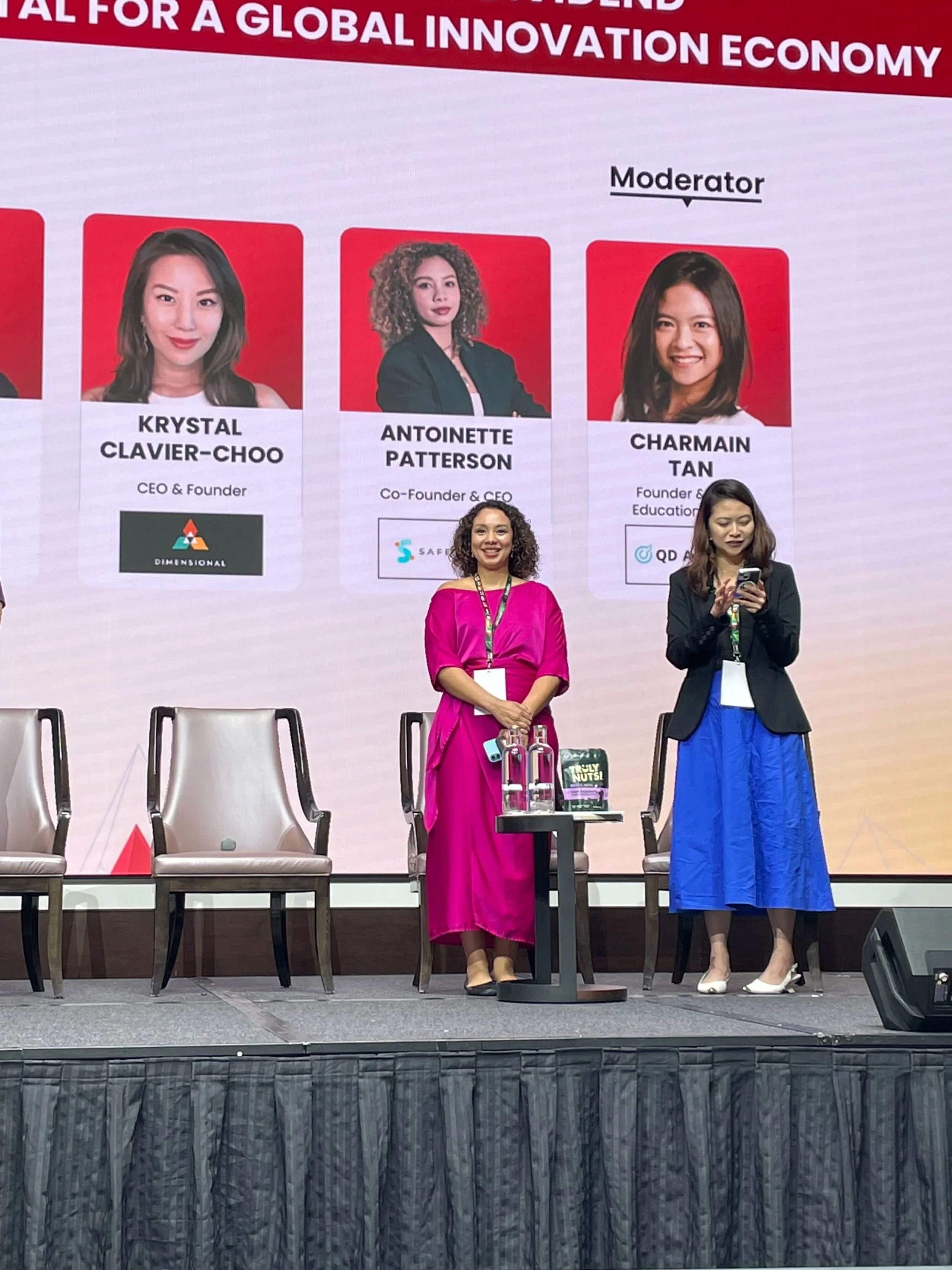 Two women are standing on a stage at a conference, with a large screen behind them displaying photos and information about three women, including their names and titles. The stage has chairs and microphones, and the women are dressed formally, one in