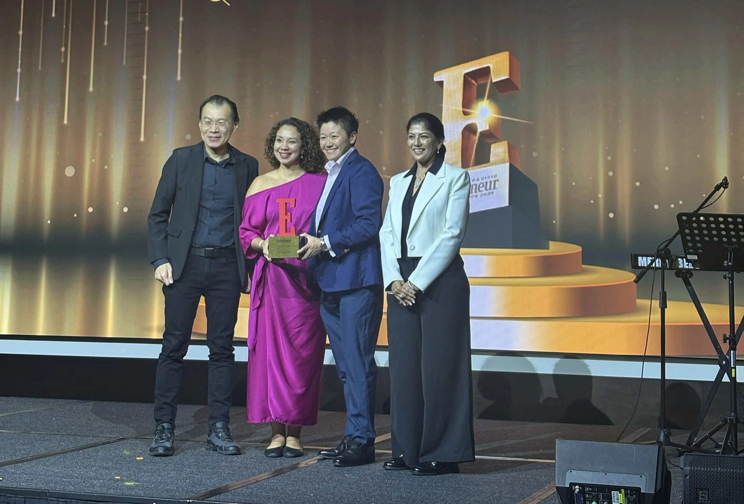 A group of four people standing on a stage at an event, with a large screen behind them displaying the letter 'E'. One woman is holding a trophy or award, and they are all smiling.
